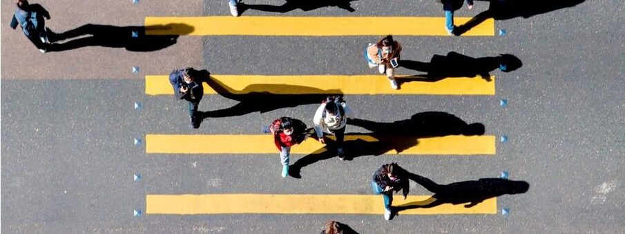 Aerial view of people crossing a street at a yellow-striped pedestrian crosswalk, casting long shadows in the sunlight.
